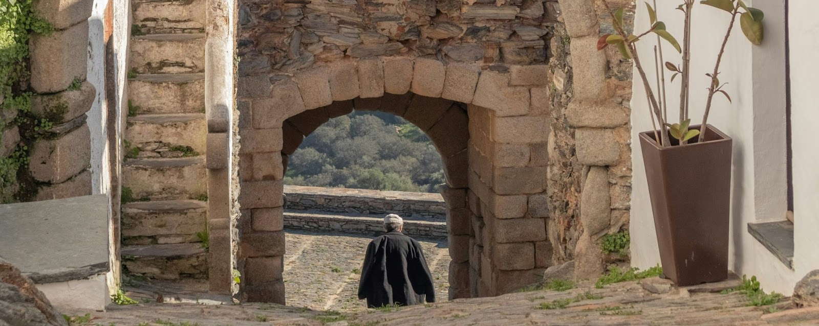 Morador caminha por rua de pedra sob arco medieval em Monsaraz, vila histórica no Alentejo; descubra onde fica Monsaraz e encante-se com seu charme rústico.
