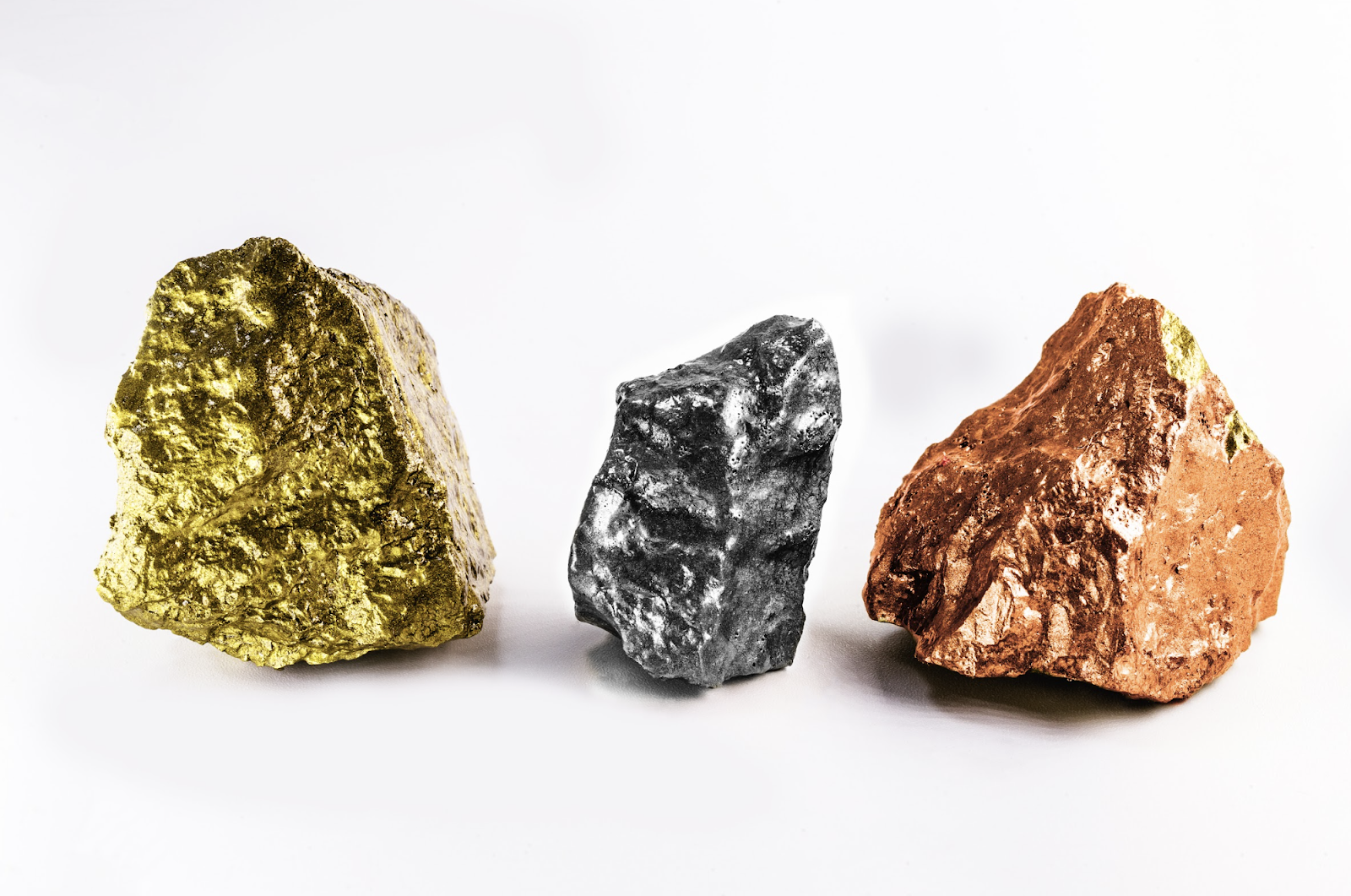 Close-up of three raw metallic rocks, gold, silver, and copper, placed on a clean white background.
