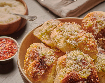 A bowl of golden-brown garlic knots sprinkled with grated cheese, served with marinara sauce and red pepper flakes on a rustic wooden table.