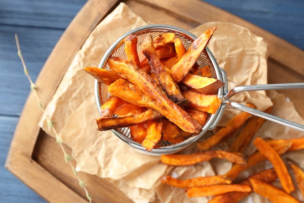 Crispy air-fried sweet potato fries served in a small metal basket atop crumpled parchment paper on a wooden board