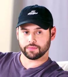 This contains an image of HYBE America CEO Scooter Braun with a beard wearing a purple shirt and a black cap looking at the camera