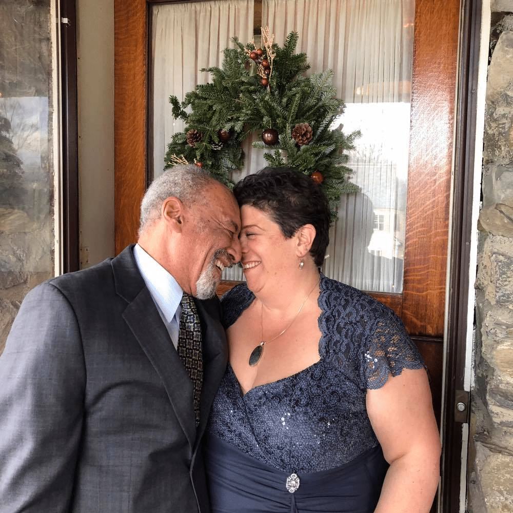 image of man in gray suit and woman in slate gray dress with foreheads together smiling at one another. They are standing in front of a door with a Christmas wreath on it.