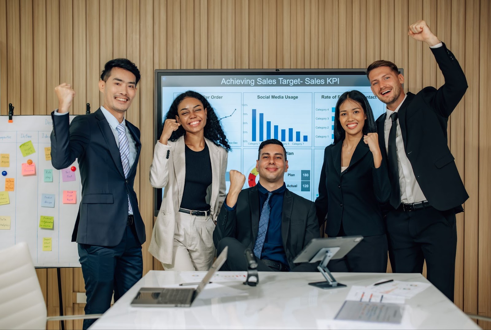 Group of men and women in business attire next to laptops and tablets with their hands in the air. 