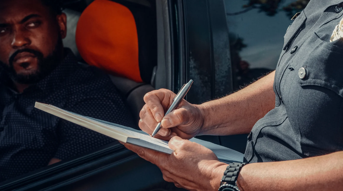 (Police officer writing a report during a traffic stop, with a driver looking on—highlighting potential unlawful citation or racial profiling.)