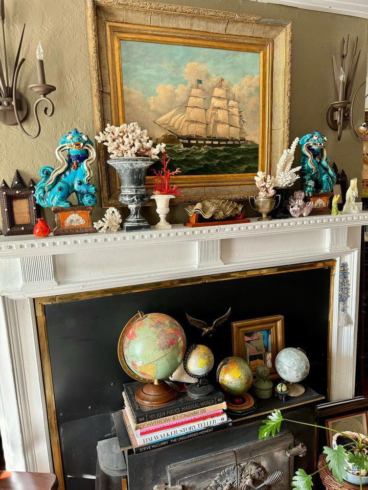 A large ship painting hangs on the fireplace with an assortment of decorative items including corals, foo dogs and globes.