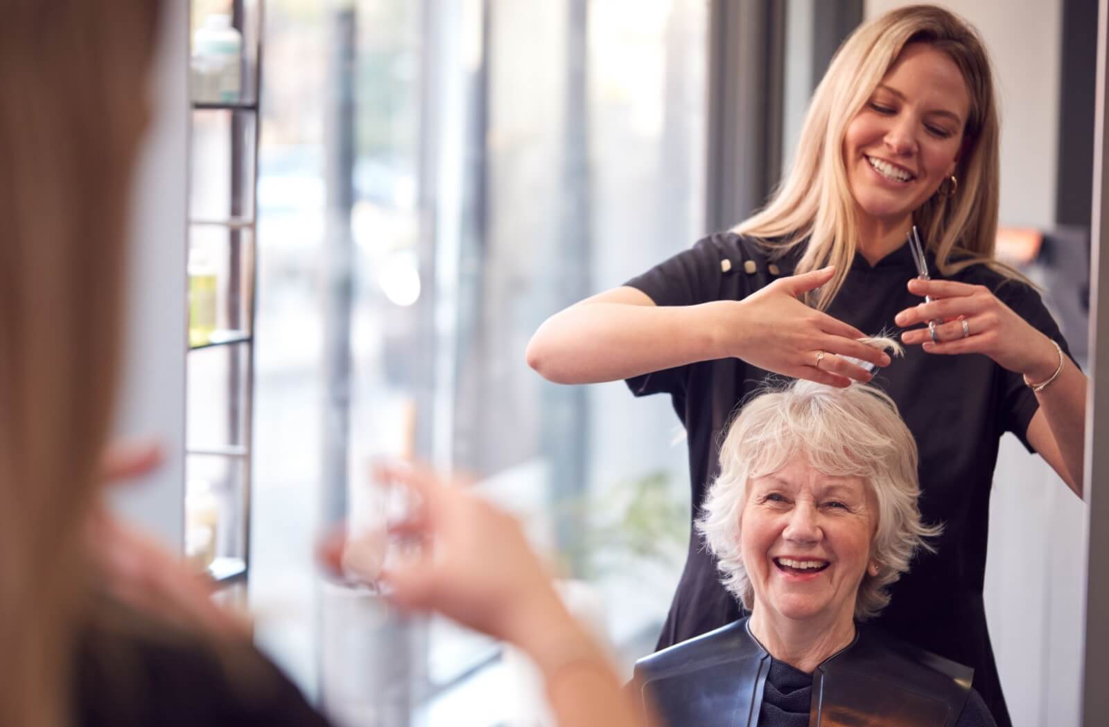 Mirror photo of a happy older adult getting their hair cut by a person with blonde hair.