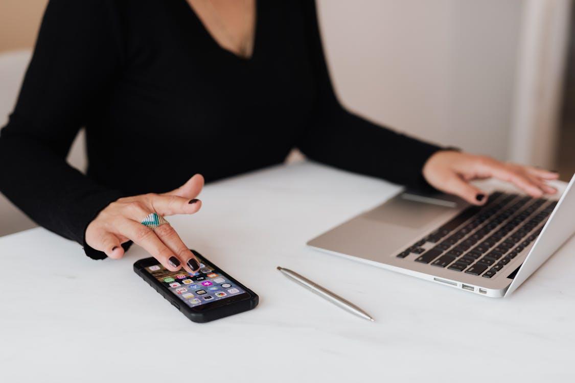 Free A professional woman multitasking with a smartphone and laptop at an office desk. Stock Photo