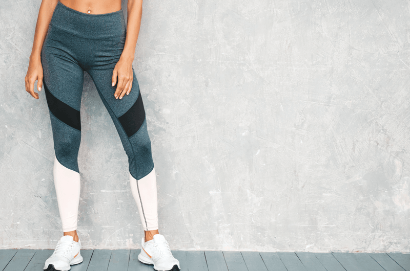 An individual is wearing athletic leggings and sneakers, standing against a textured gray wall. The leggings are primarily dark gray with black diagonal stripes on the thighs and light gray sections on the lower legs. The person is standing on a wooden floor painted in a light blue-gray color.