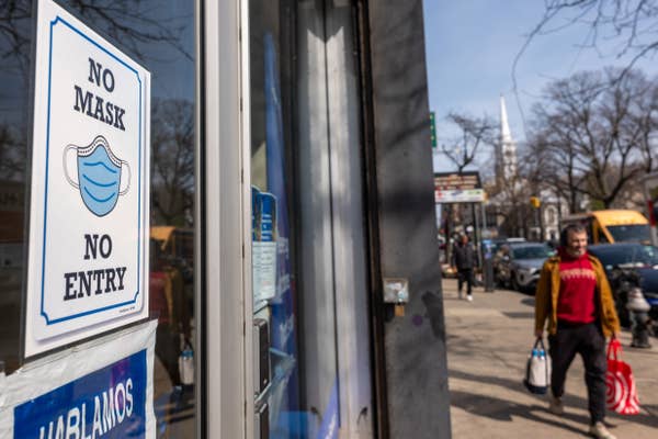 Sign on store door reads &quot;No Mask, No Entry&quot; with a face mask icon. A person carrying shopping bags walks by on a busy street