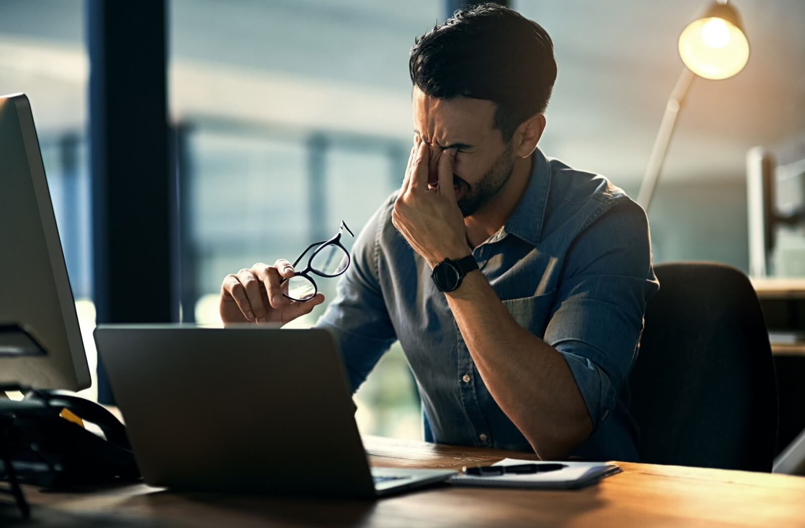 A person with eye strain rubs their eyes while sitting in front of multiple computer screens in a dimly lit office.