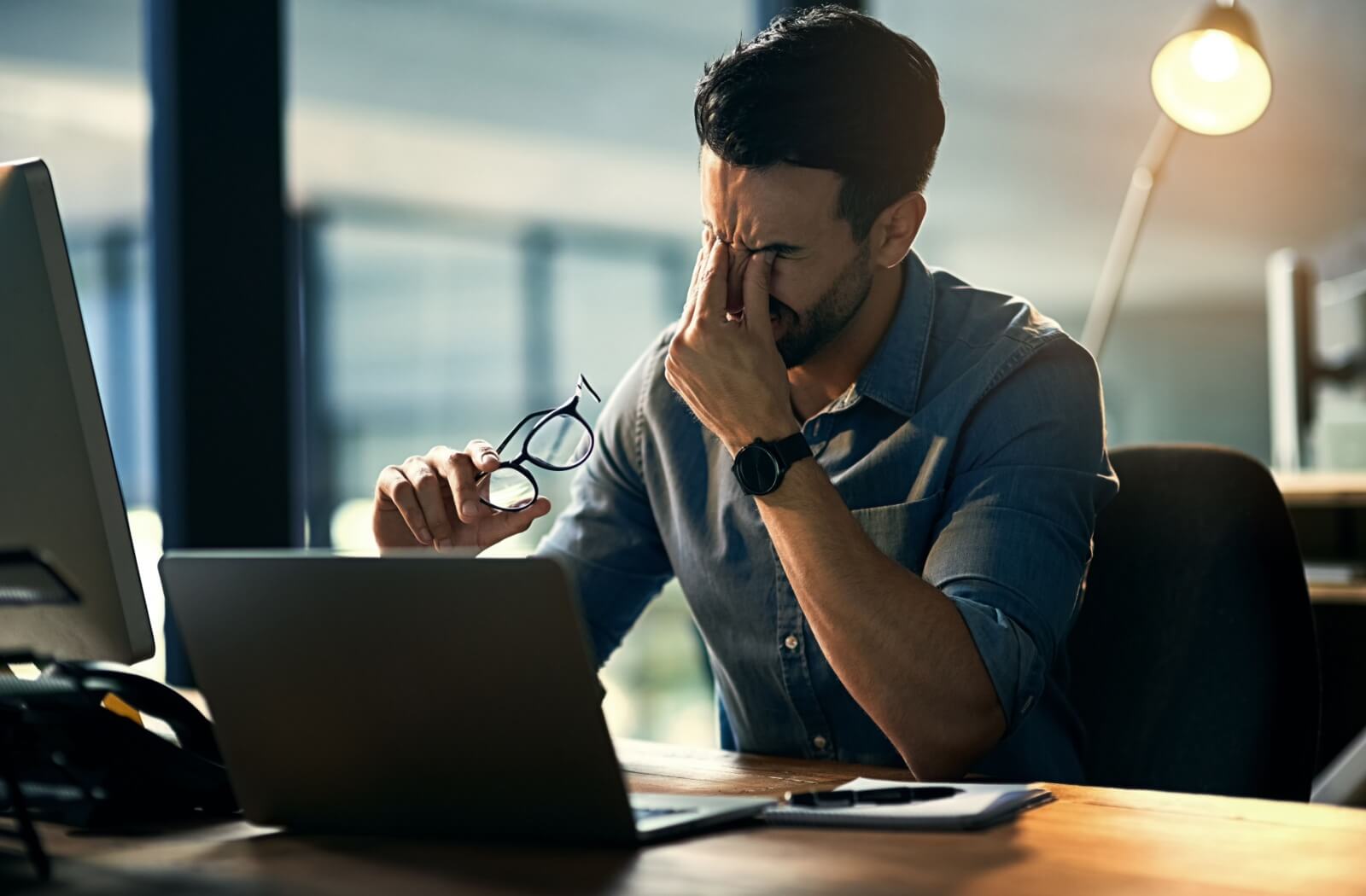 A person with eye strain rubs their eyes while sitting in front of multiple computer screens in a dimly lit office.