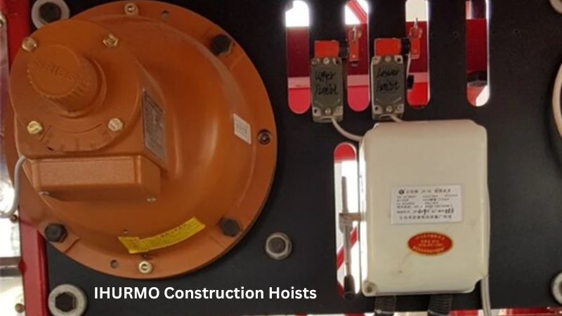 Close-up dos controles do elevador de construção IHURMO: peças elétricas etiquetadas e motor laranja em um painel.