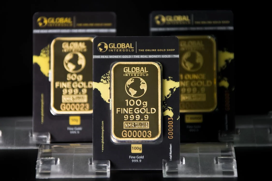 Three gold bullion bars in their protective packaging on a counter, including a 100-gram bar, a 50-gram bar, and an unidentified bar, symbolizing investment in precious metals.