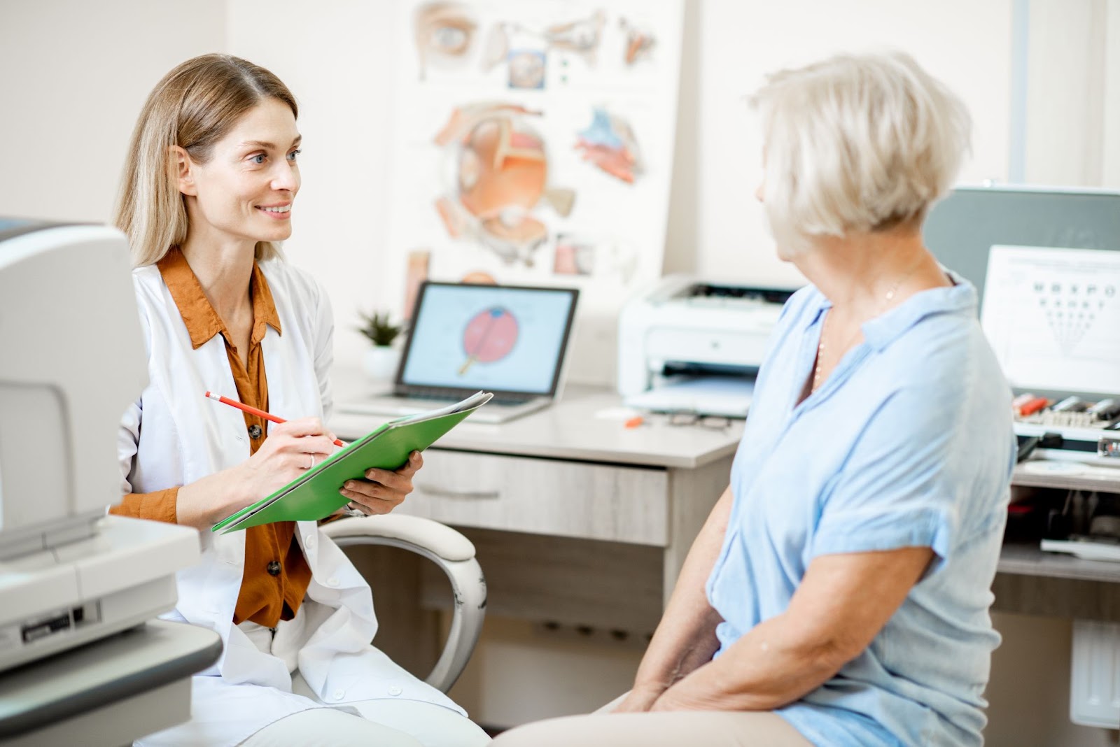 An optometrist sitting in her office surrounded by optometric equipment as she writes on her green notepad while smiling and speaking to her older adult patient.