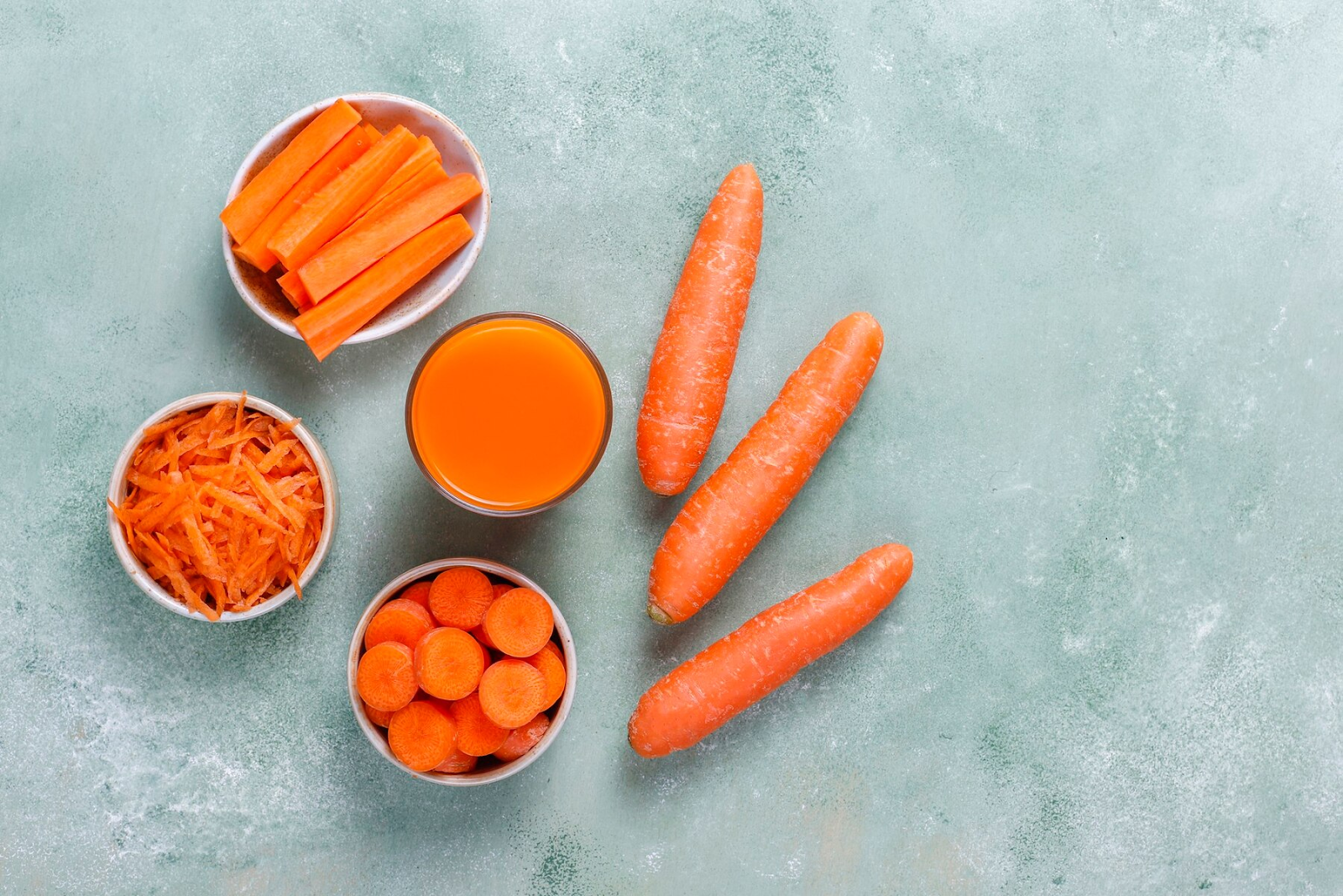 Three whole carrots, carrot juice, and differently-sliced carrots in separate bowls