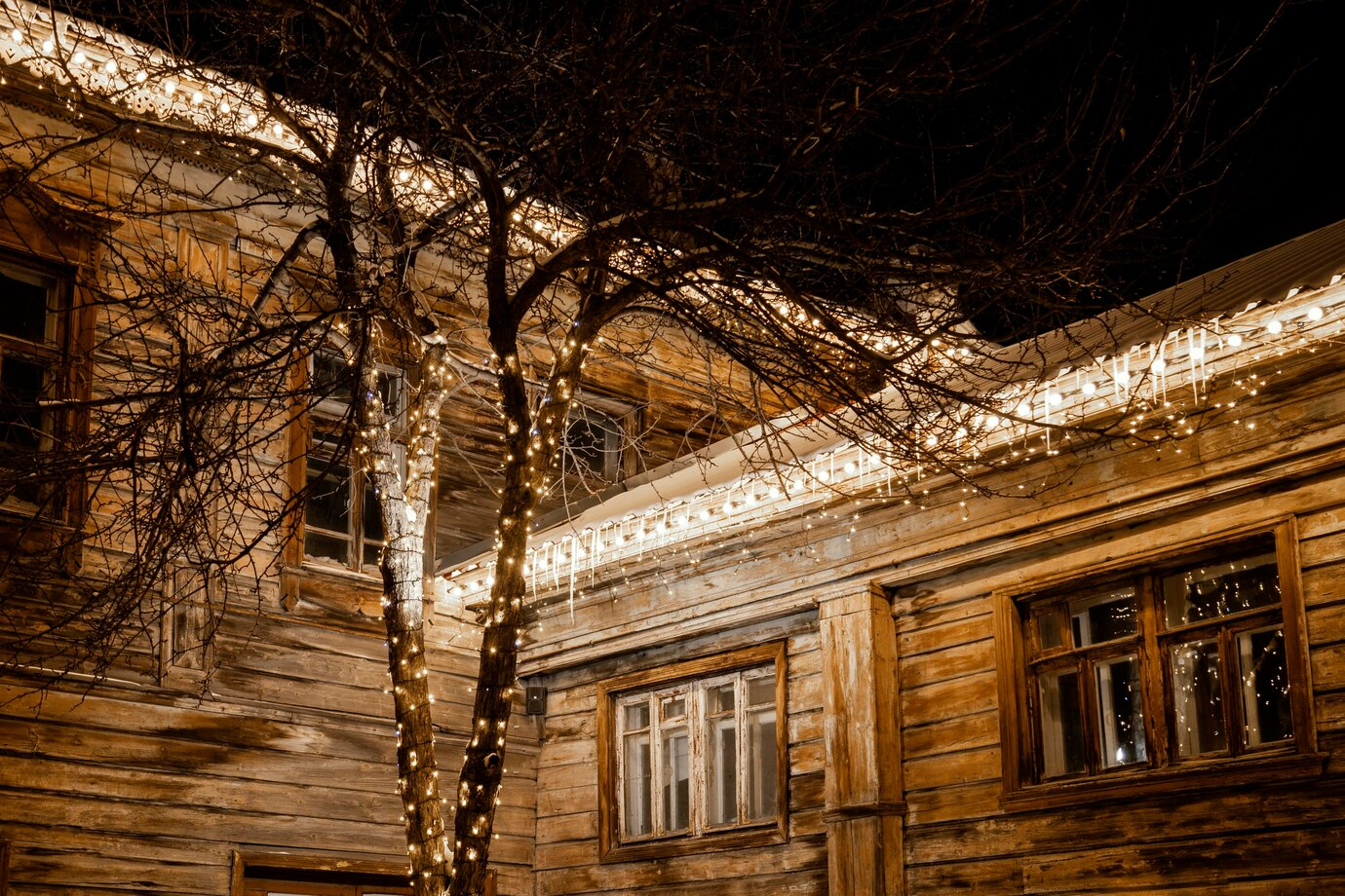 A rustic wooden house adorned with warm string lights along the roofline and wrapped around the bare branches of nearby trees, creating a festive and cozy ambiance against the dark night sky