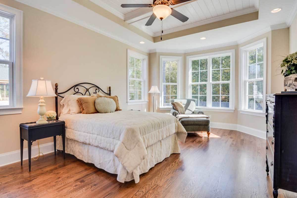 Primary bedroom with a tray ceiling, beige walls, bay window, and an ornate bed complemented with dark wood nightstands and a cozy chaise lounge.