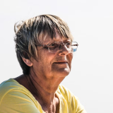 A person with short hair and glasses, wearing a yellow shirt, looks thoughtfully to the distance.