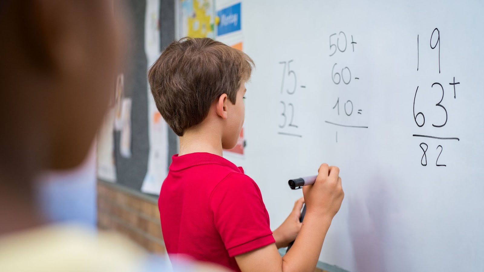 A young boy in a red shirt writing mathematical problems on a whiteboard in a classroom, representing preparation for dyscalculia testing.