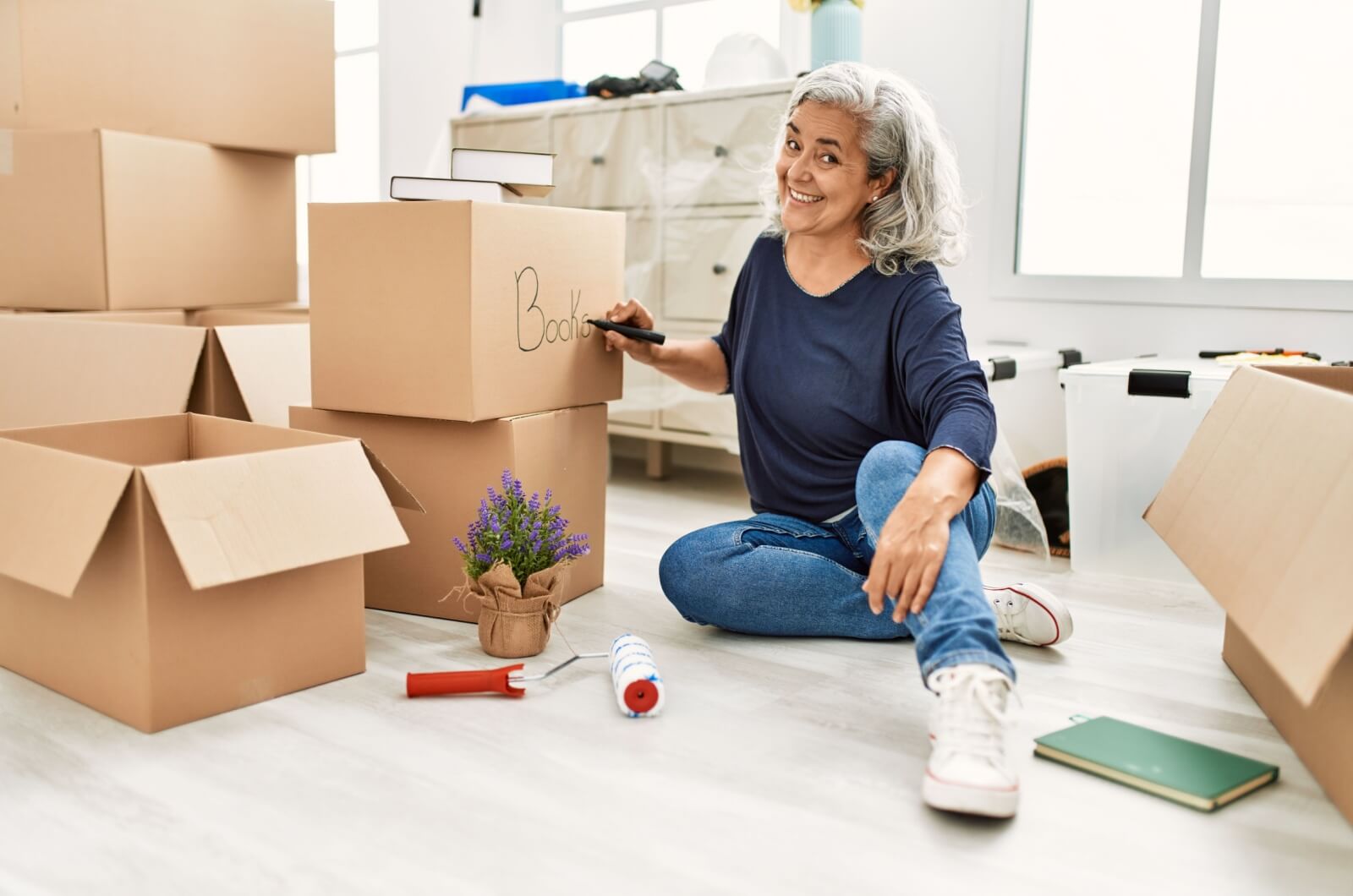 A smiling older adult sitting on the floor labelling a box with books inside.