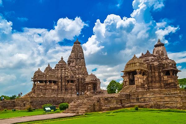 A stone building with a green lawn and blue sky with Khajuraho Group of Monuments in the background

Description automatically generated