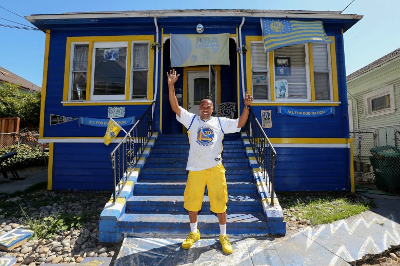 A person in yellow shoes, yellow shorts and a white Golden State Warriors basketball jersey stands with their arms in the air in front of a blue and yellow home, with Golden State Warriors memorabilia adorning the exterior.