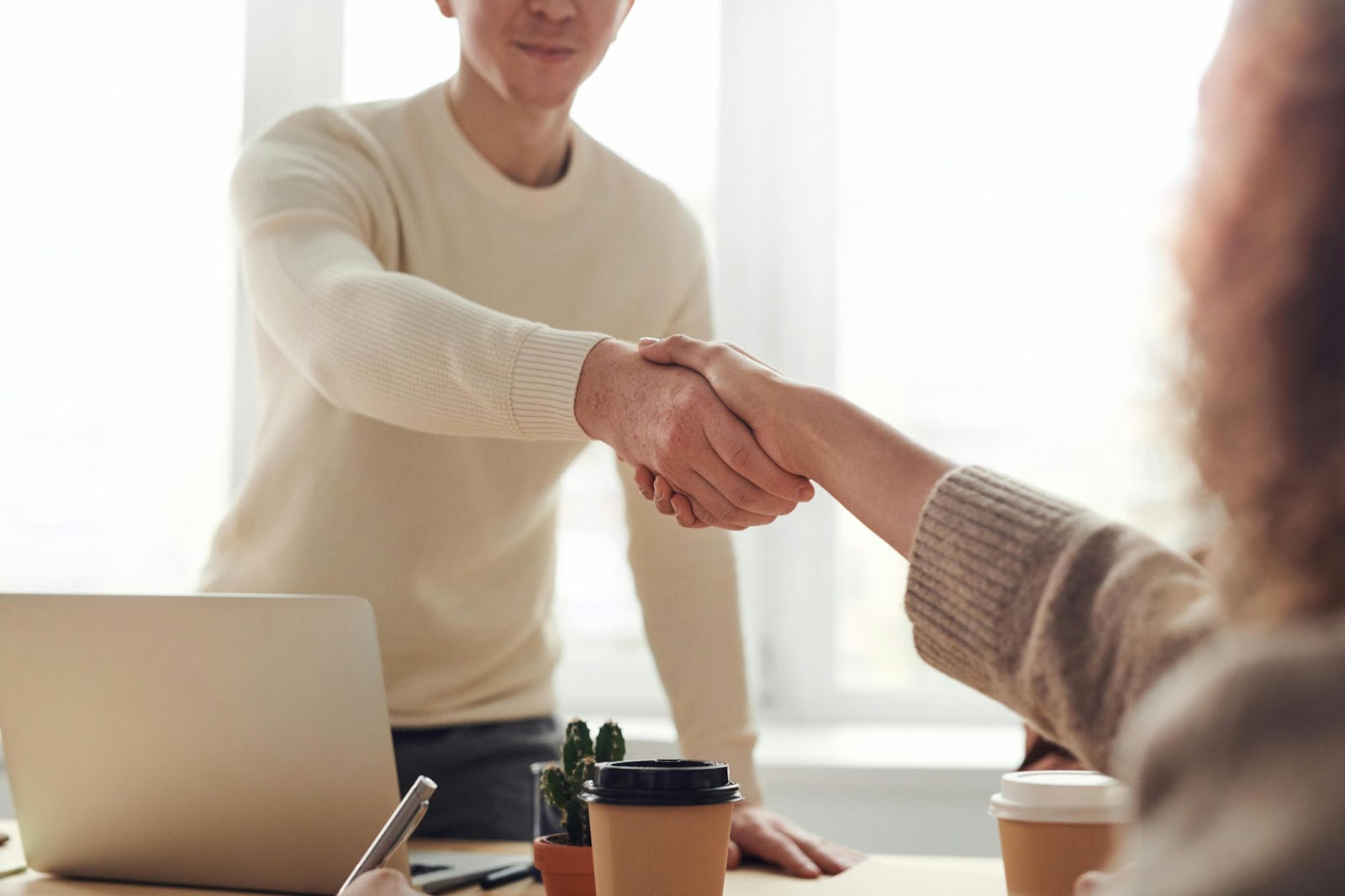 A young man in a white tee shakes hands with a female colleague in an office, with a laptop and coffee mug on the table.