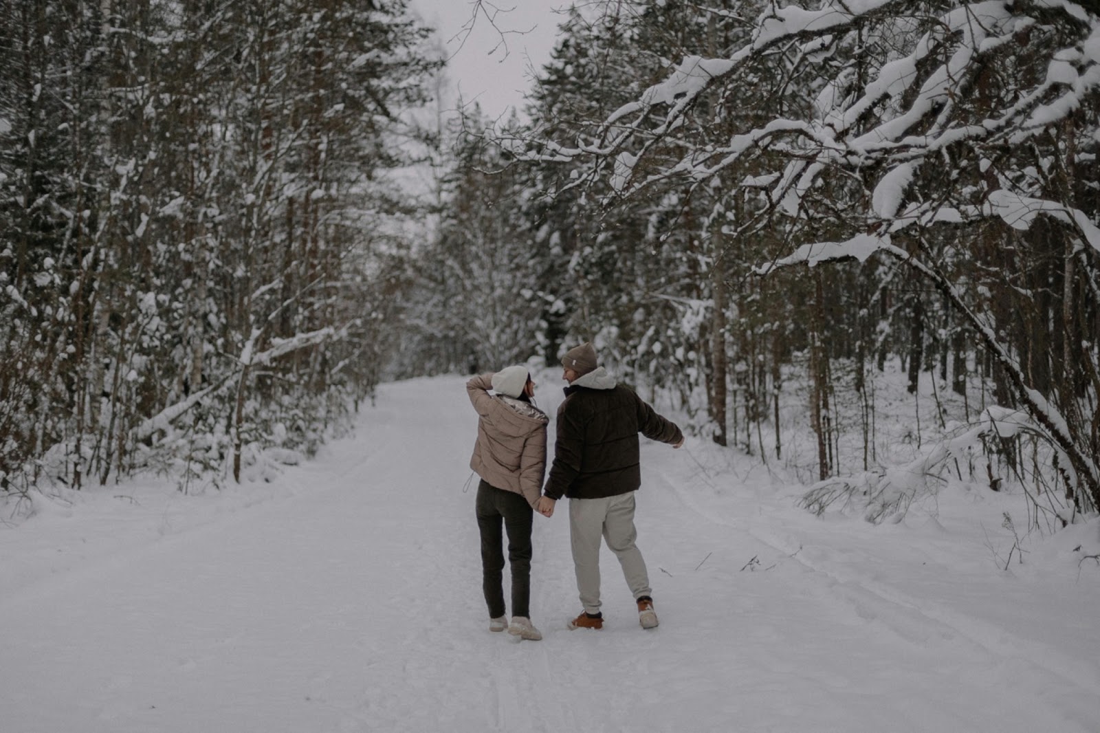 A man in a black jacket, holding the hand of a woman. While they are both wearing a hood, they both look at each other with a happy expression. They are in a beautiful winter landscape, in a forest full of snow.