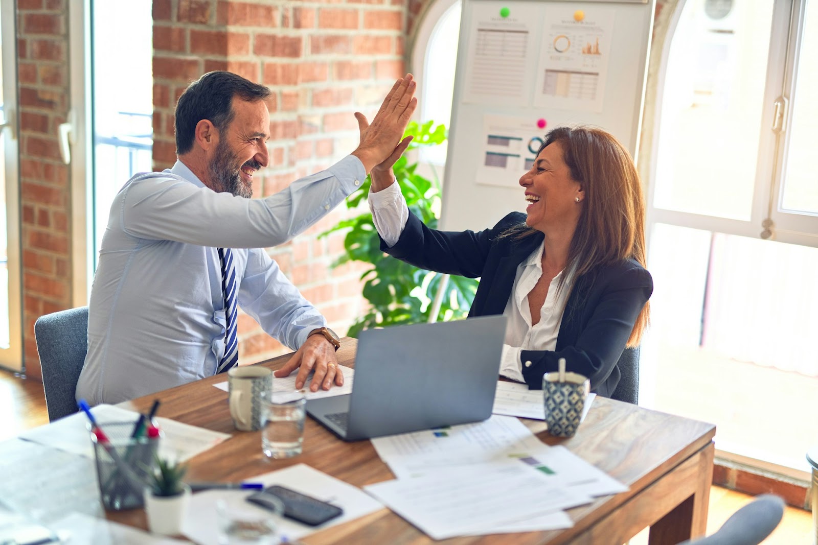 Two business professionals giving each other a high five at a desk, celebrating a successful collaboration or achievement.