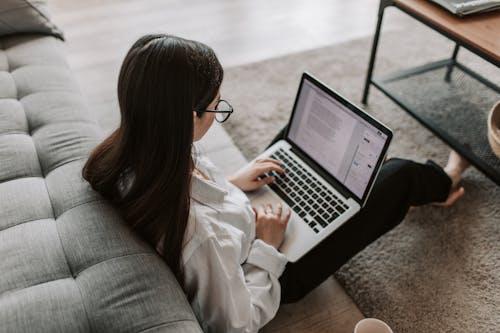 Free Woman working remotely with a laptop on the floor next to a sofa, enjoying comfortable home office setup. Stock Photo