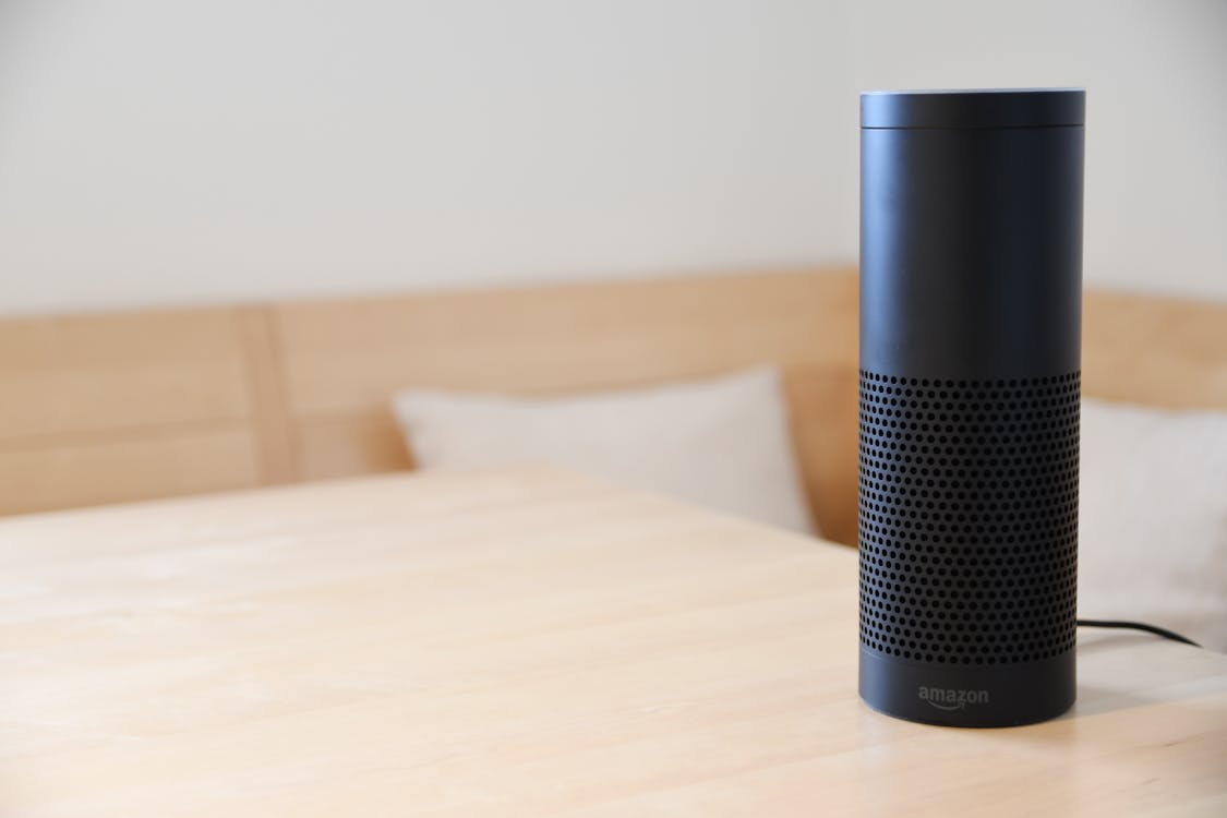 Free A black smart speaker resting on a light-colored wooden table in a cozy indoor setting. Stock Photo