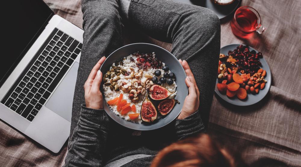 Una persona sosteniendo un plato de frutas, verduras y frutos secos junto a su laptop