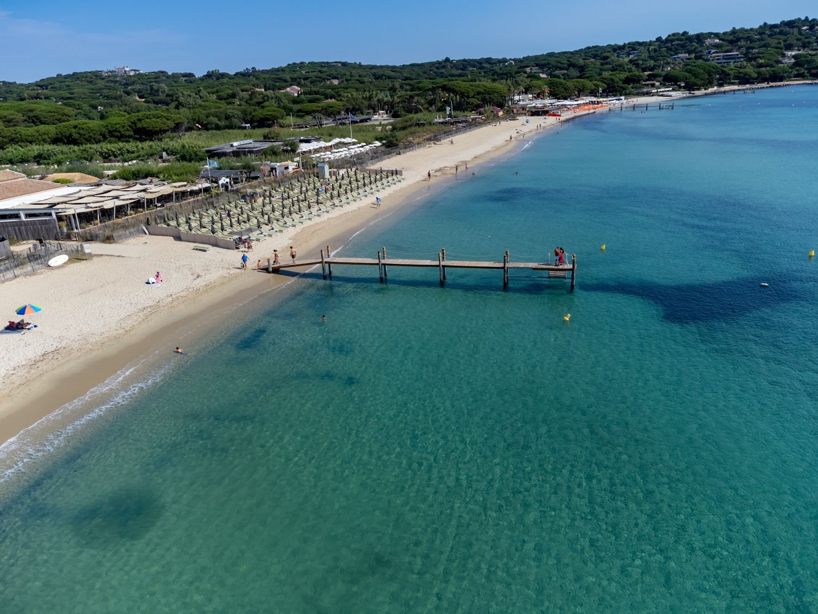 Aerial view of Pampelonne Beach near Saint-Tropez, France, showing clear turquoise water, soft sandy shoreline, a wooden pier and rows of sun loungers backed by green vegetation.