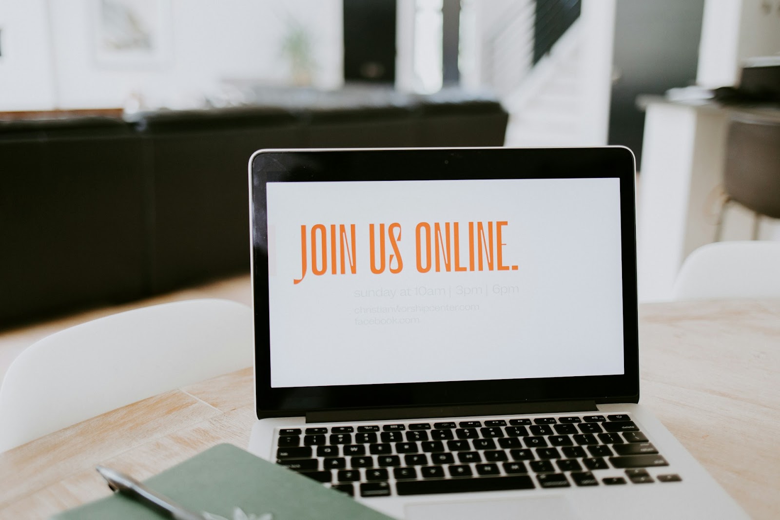 A laptop on a wooden table displays an invitation to 'Join Us Online' for virtual gatherings, listing times and social media links, in a stylish modern interior.