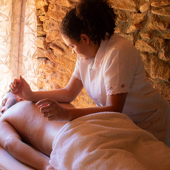 Uma profissional de massagem realizando massagem relaxante em uma pessoa deitada, com fundo de parede de pedra e cortina de madeira, promovendo bem-estar e relaxamento.