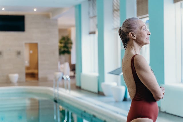 Older woman standing outside of a an indoor swimming pool, looking out of a window.