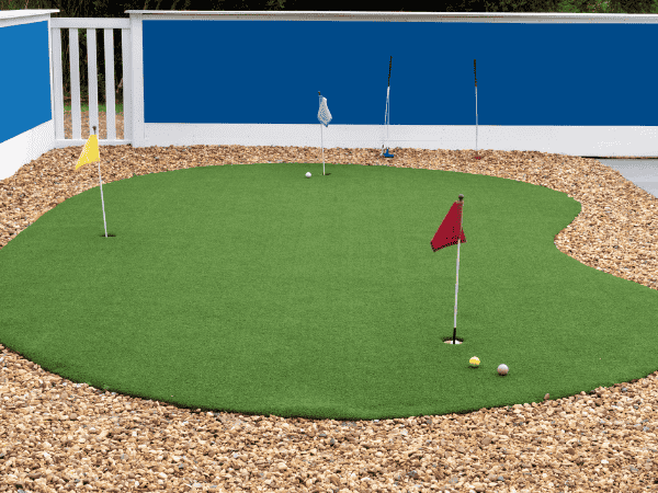 Backyard artificial turf putting green with three flagsticks and golf balls surrounded by stone mulch