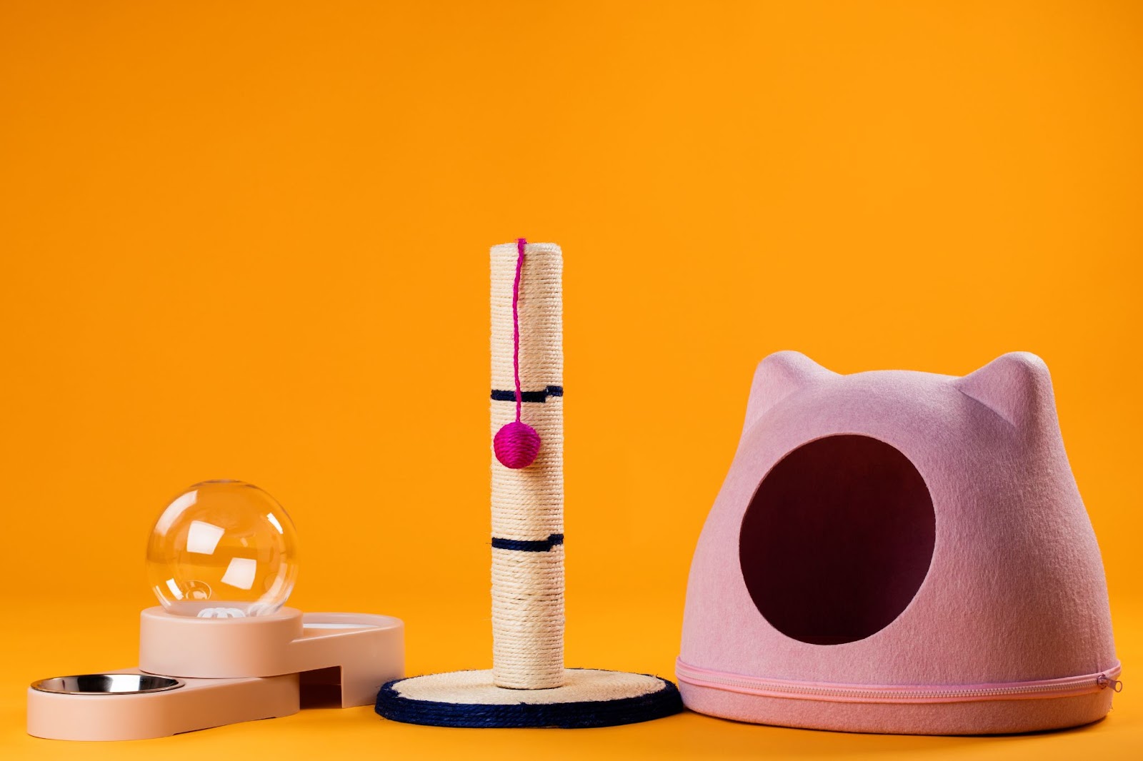 A set of cat supplies for a new cat, featuring a scratching post with a pink toy, a modern cat house, and food and water bowls, all against a bright orange background.