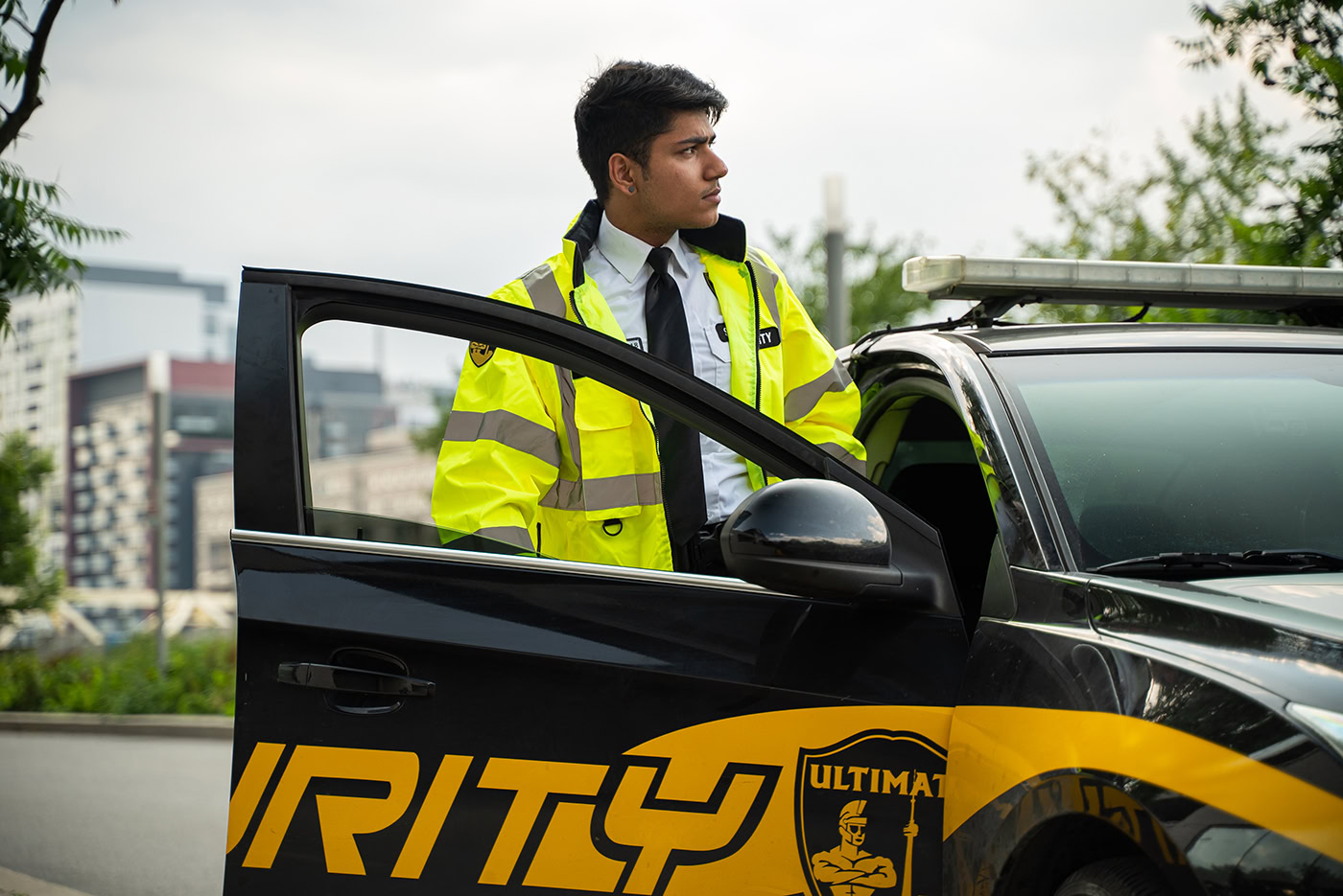 A security guard in a yellow jacket, who had to obtain a security license, stands next to a black security vehicle with the driver’s door open in an urban setting.