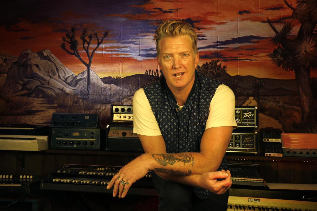 Josh Homme leaning on a soundboard in a studio wearing a white T-shirt and black vest