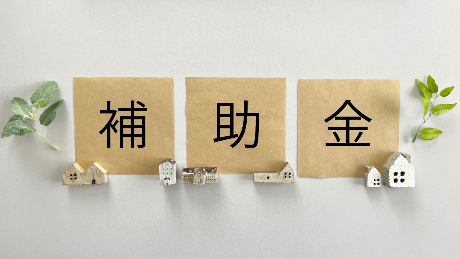 「補助金」と書かれた3枚の茶色い紙と、複数のミニチュアの家、そして緑の葉の飾り