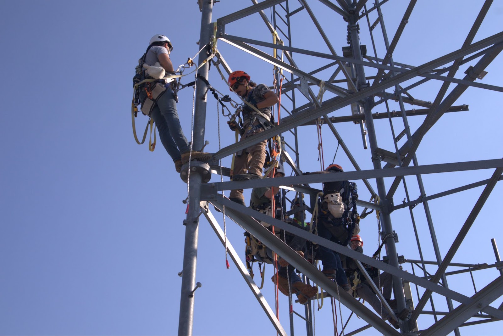 A group of telecommunications tower technicians wearing safety harnesses and helmets work together on a steel tower structure. One technician stands on a beam, observing or guiding others, while three technicians are positioned on different parts of the structure, engaging with their equipment. The clear blue sky provides a contrasting backdrop, emphasizing the height and structure of the tower.