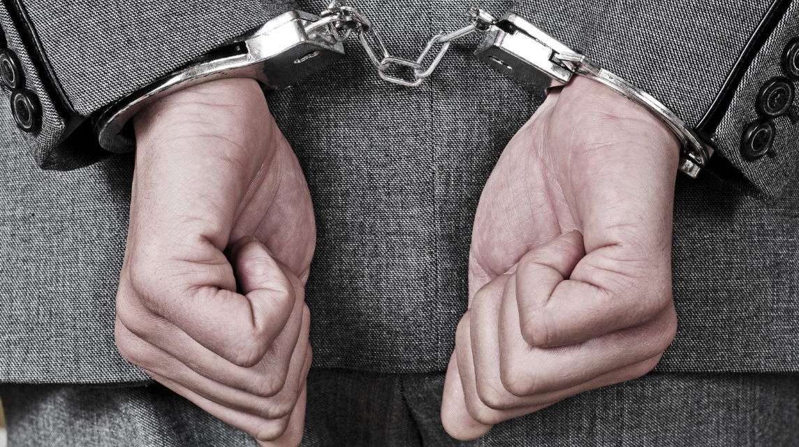(Close-up of a person in a suit wearing handcuffs, representing wrongful arrest or civil rights violations related to unlawful detainment.)