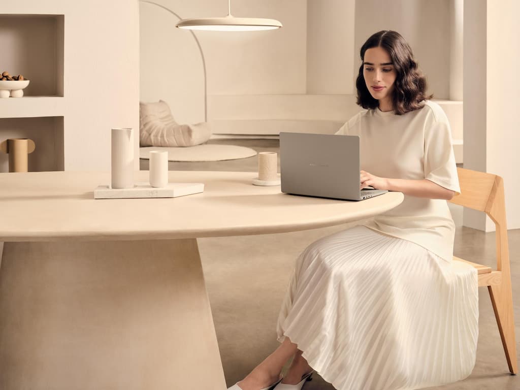 Uma mulher, vestida de branco, trabalhando em seu Zenbook A14 apoiado em uma mesa bege redonda, em um ambiente moderno e com tons claros.  