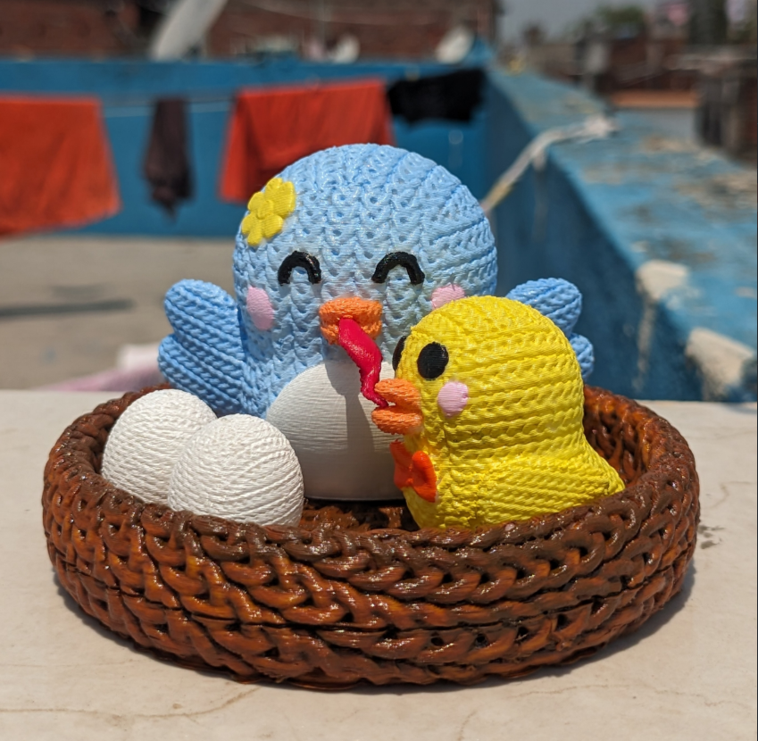 Crochet-style 3D printed mama bird and yellow baby chick sharing a worm in a woven nest with white eggs, designed by Anukalpgenesis3d

