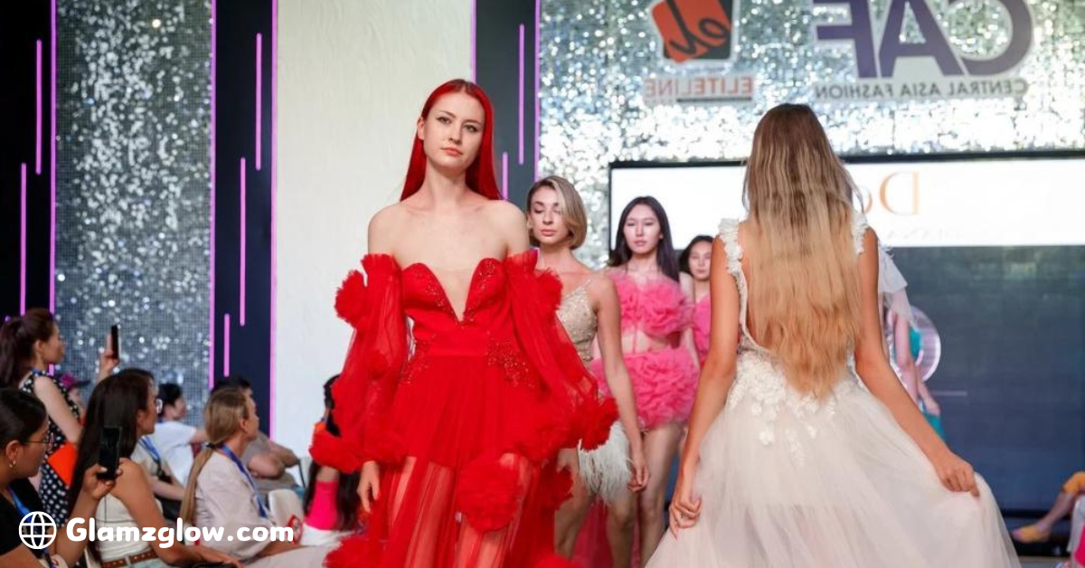 Models walking the runway during a fashion show, featuring a lead model in a striking red dress, followed by others in pastel-colored gowns, with an audience watching closely.