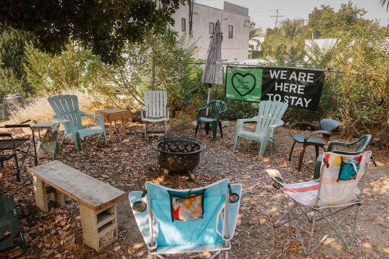 An array of lawn chairs and and wooden bench are set in a circle around a fire pit. The ground is littered with leaves and the area is surrounded by vegetation and a chain-linked fence. A sign on the fence reads, "We Are Here to Stay."