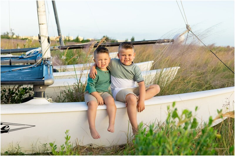 Two young boys smiling and sitting on a white sailboat, captured by a Love and Style photographer on Anna Maria Island.