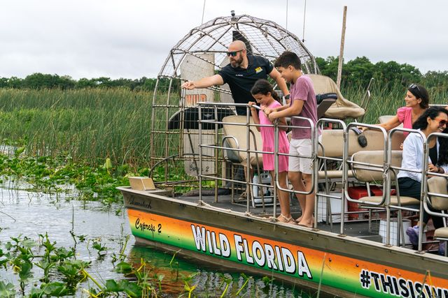 A group of people enjoying a Wild Florida airboat tour, with a guide pointing out to the water as children look on.