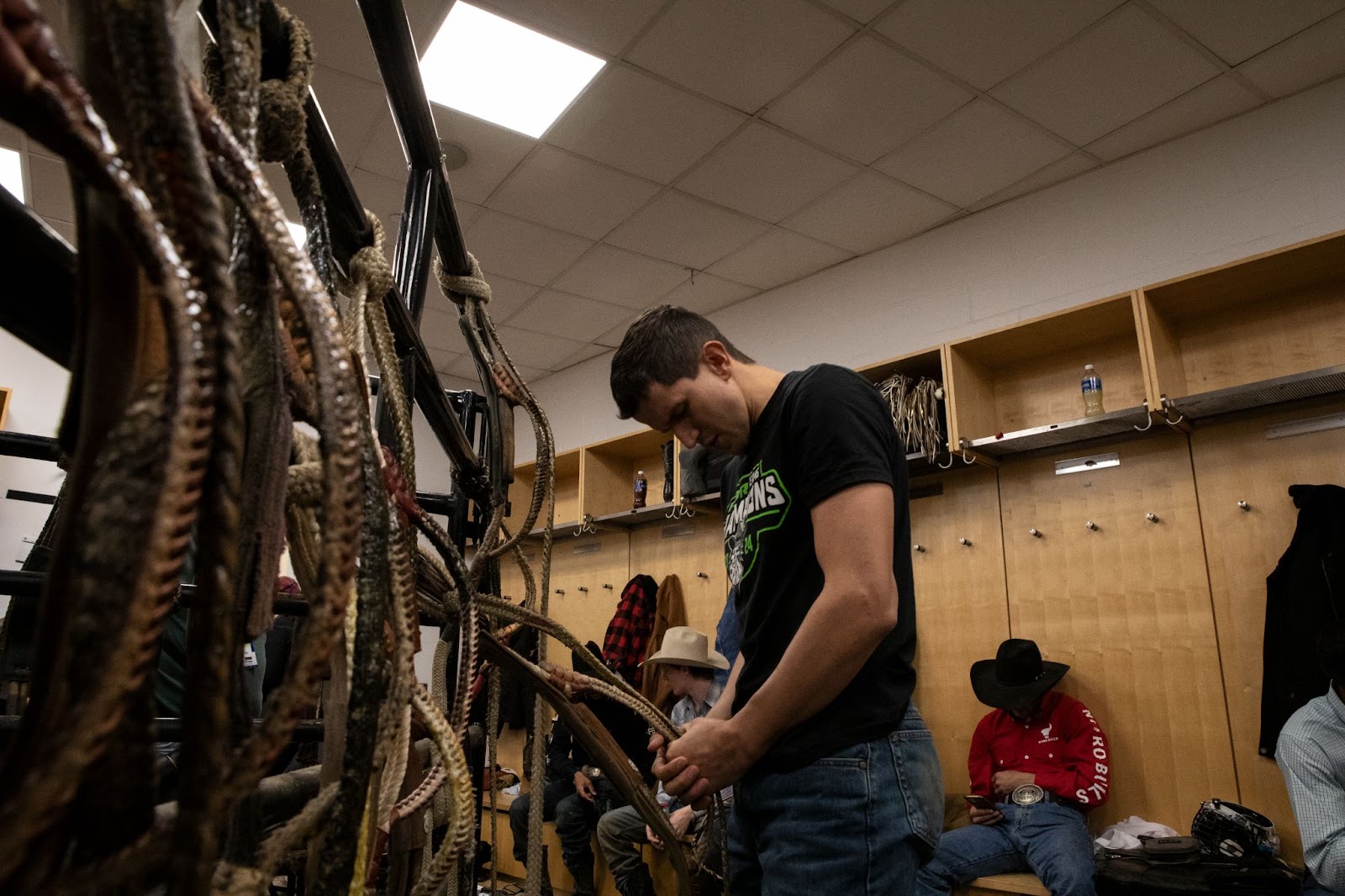 Rider wearing black T-shirt and jeans prepares rope in locker room before the event begins.